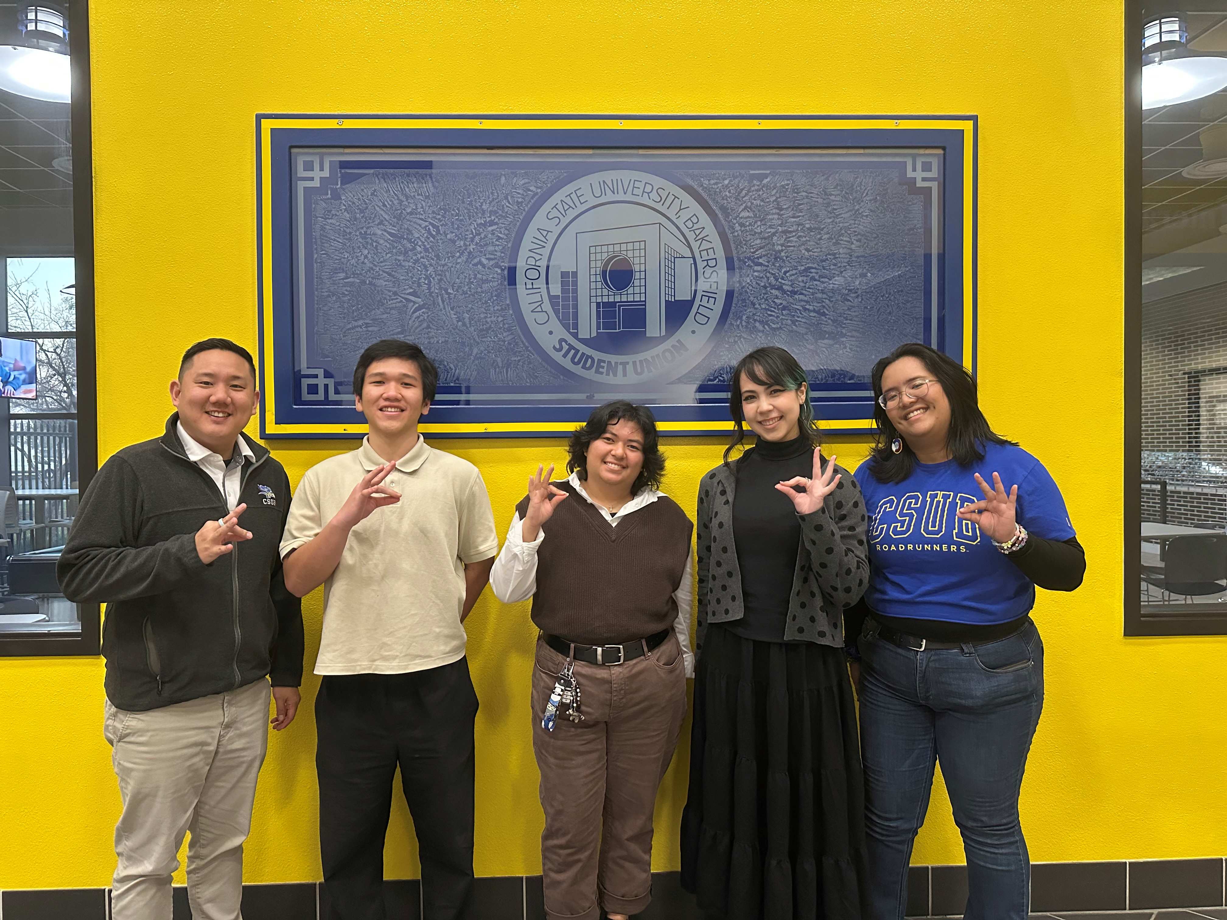 5 people stand in front of CSUB logo, putting up the runner hand signal of an "OK" sign with a pointed beak. This picture from left to right is Mike Kwon, Duong Nguyen Long Nguyen, Gabrielle Becina, Alexia Nankil, and Marrian Angeles.