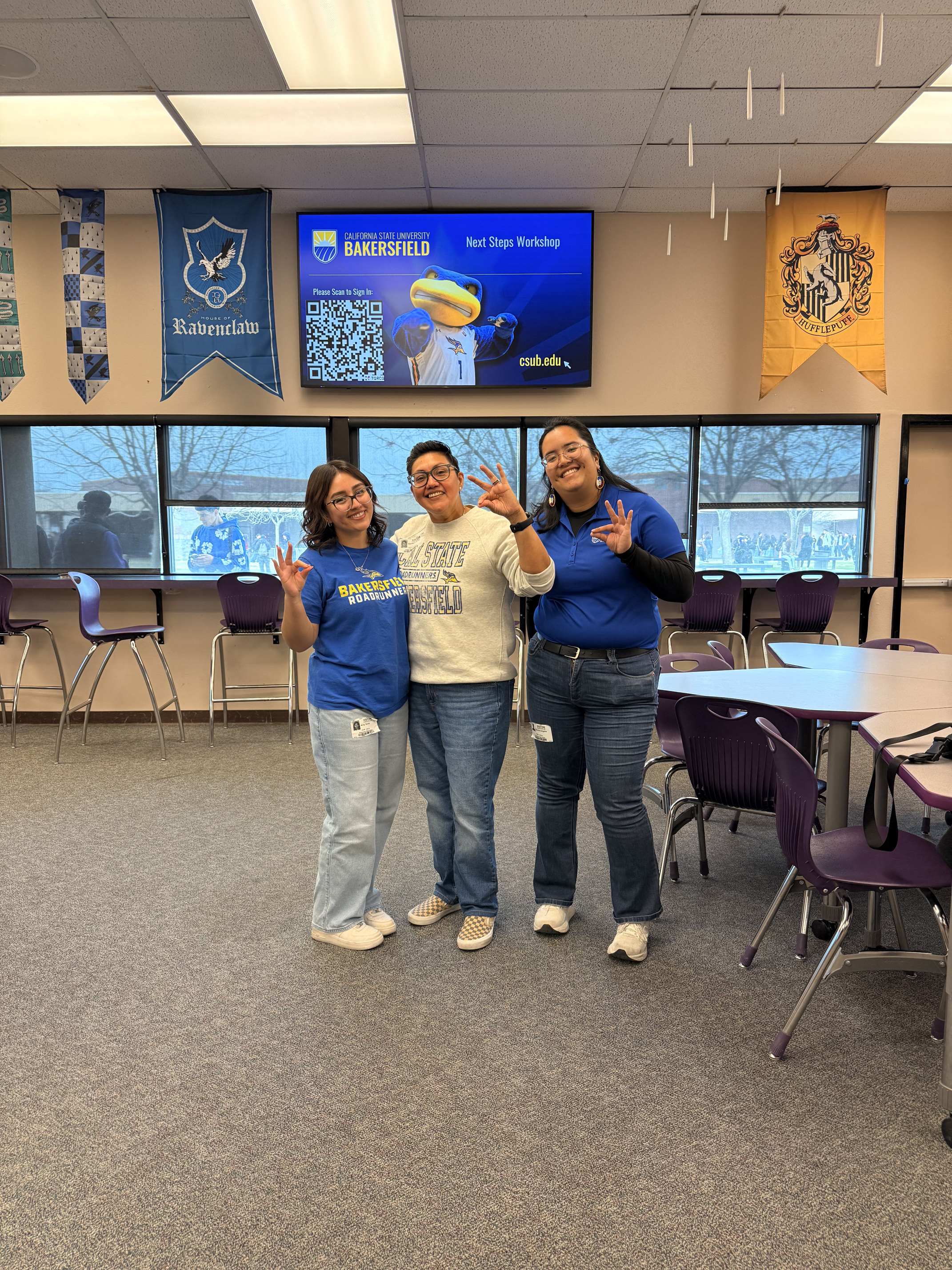 Three people with blue and white shirts, holding up an OK sign with a pointed beak. They are standing in front of a TV stating "CSU Bakersfield - Next Steps Workshop" with a Roadrunner mascot, QR Code, and the website "csub.edu" on it.